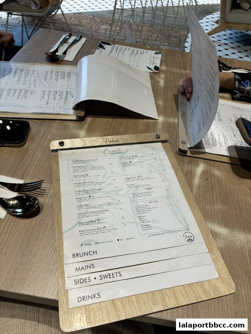 A menu from POKOK LaLaport is open on a wooden table with cutlery.