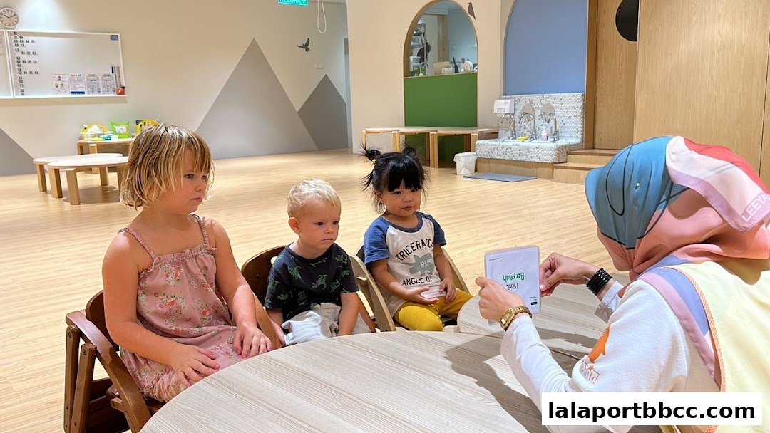 A teacher shows a card to three young children sitting at a table in a bright classroom.