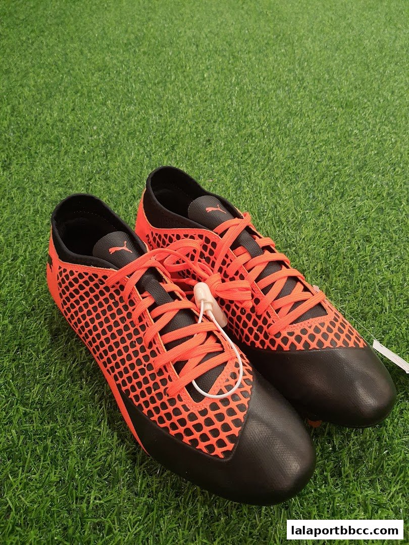 A pair of bright orange and black Puma soccer cleats on a green artificial turf.