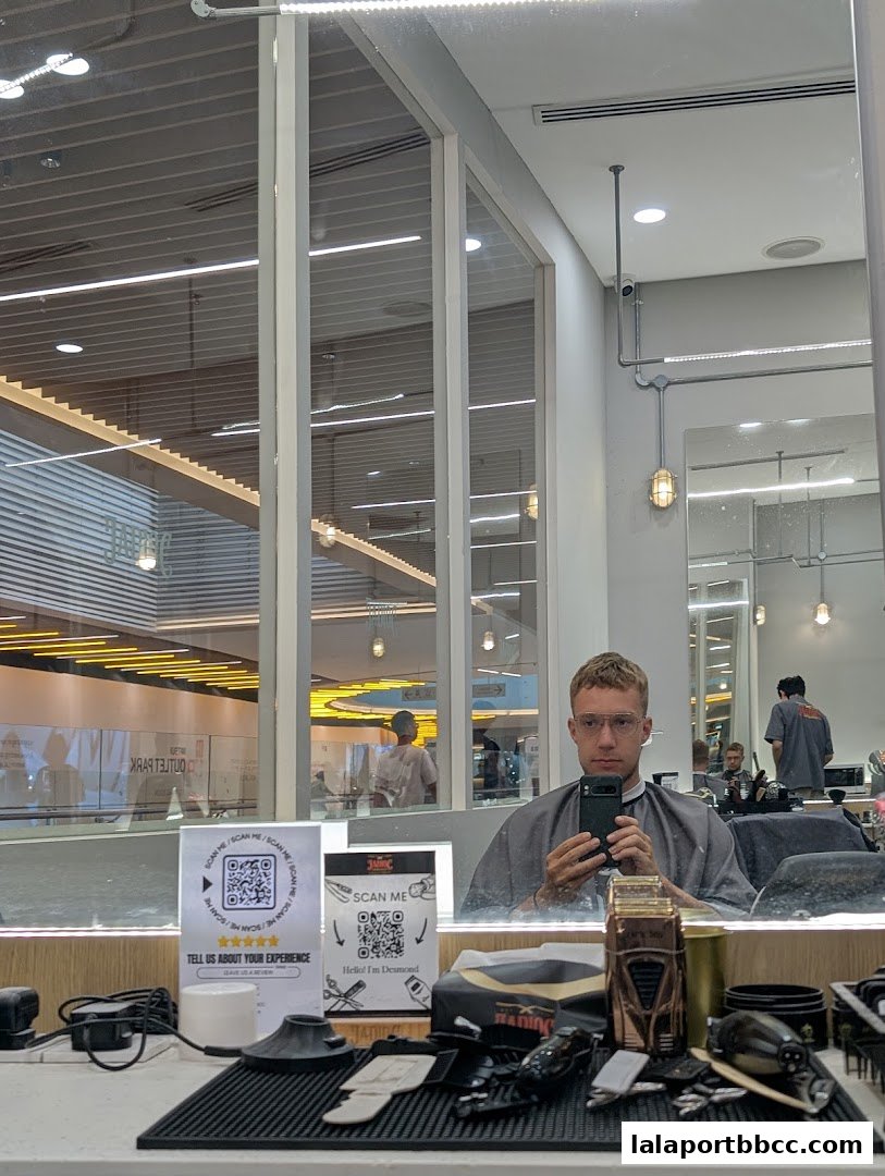 A man in a barber cape takes a selfie in a modern barbershop, with reflections of the mall visible.