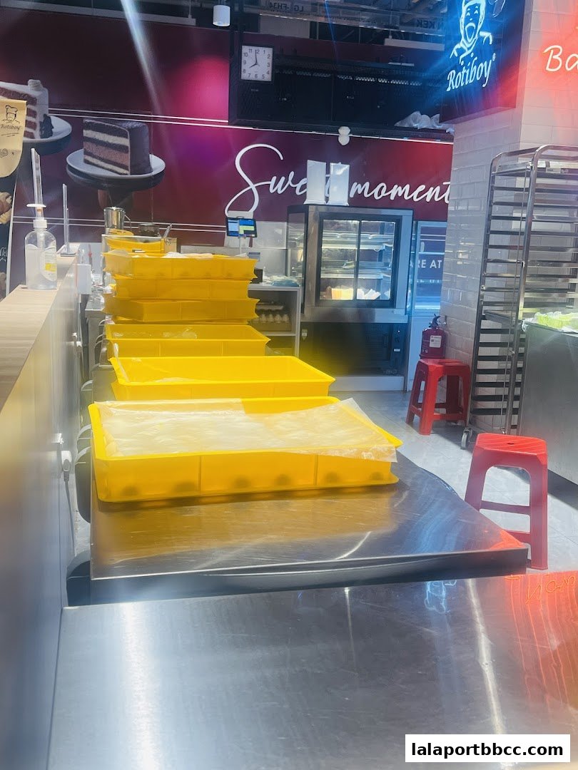 A bakery counter with yellow trays filled with baked goods, a display case, and Roti Boy branding.