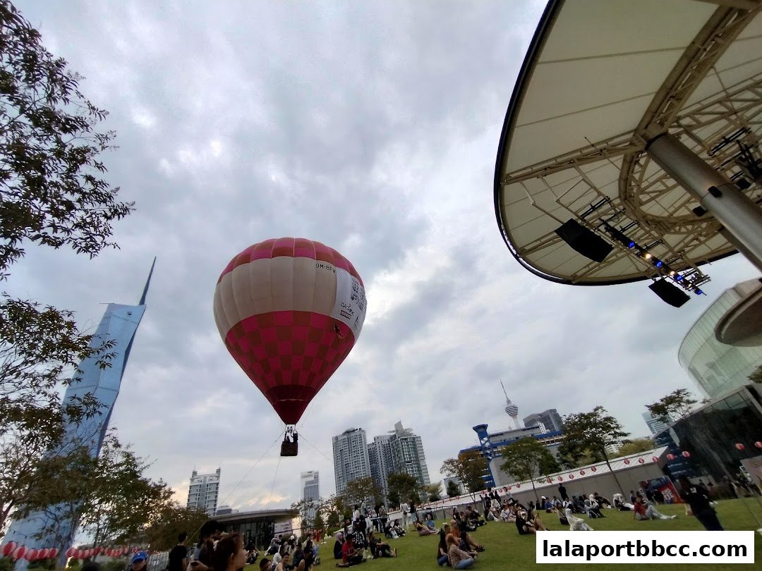 A hot air balloon floats over a grassy area with people sitting and a modern building in the background.