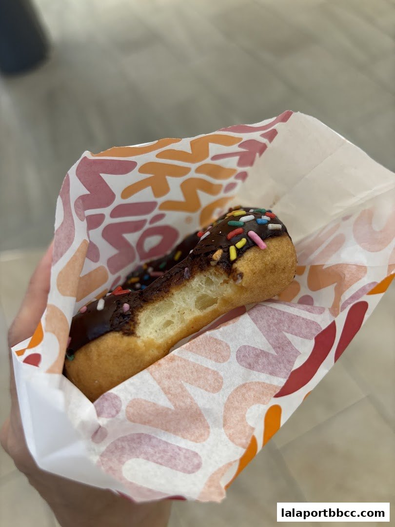 A hand holds a Dunkin' Donuts bag with a chocolate-frosted donut with sprinkles.