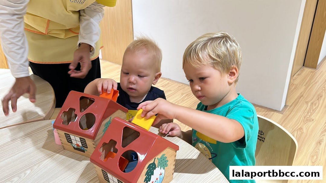 Two young children are playing with shape sorter toys at a table in a brightly lit room.
