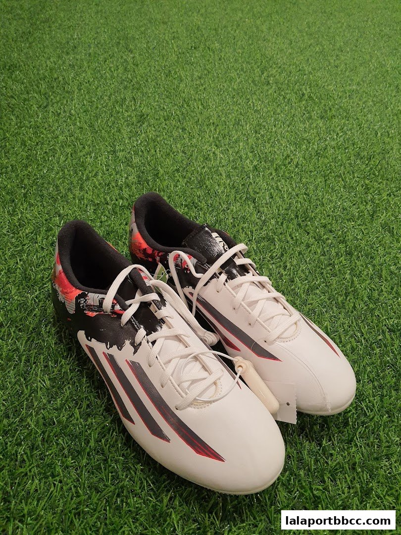 A pair of white Adidas soccer cleats with red and black accents on a green turf background.