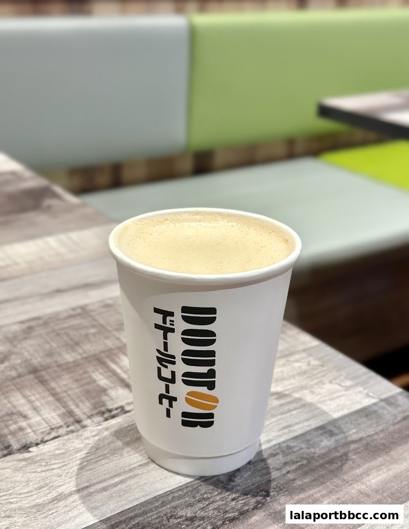 A white paper cup with the DOUTOR COFFEE logo and Japanese text sits on a wooden table.