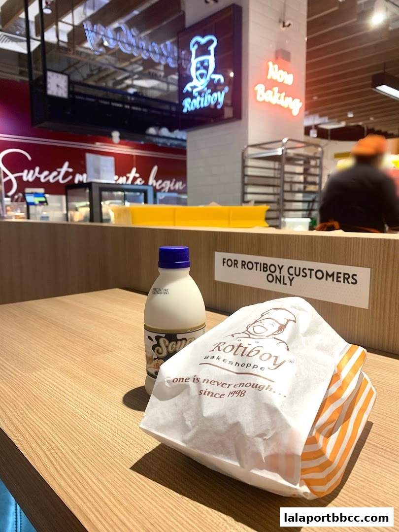 A Roti Boy paper bag and a bottle of drink on a wooden counter with the shop's logo and 'Now Baking' sign.