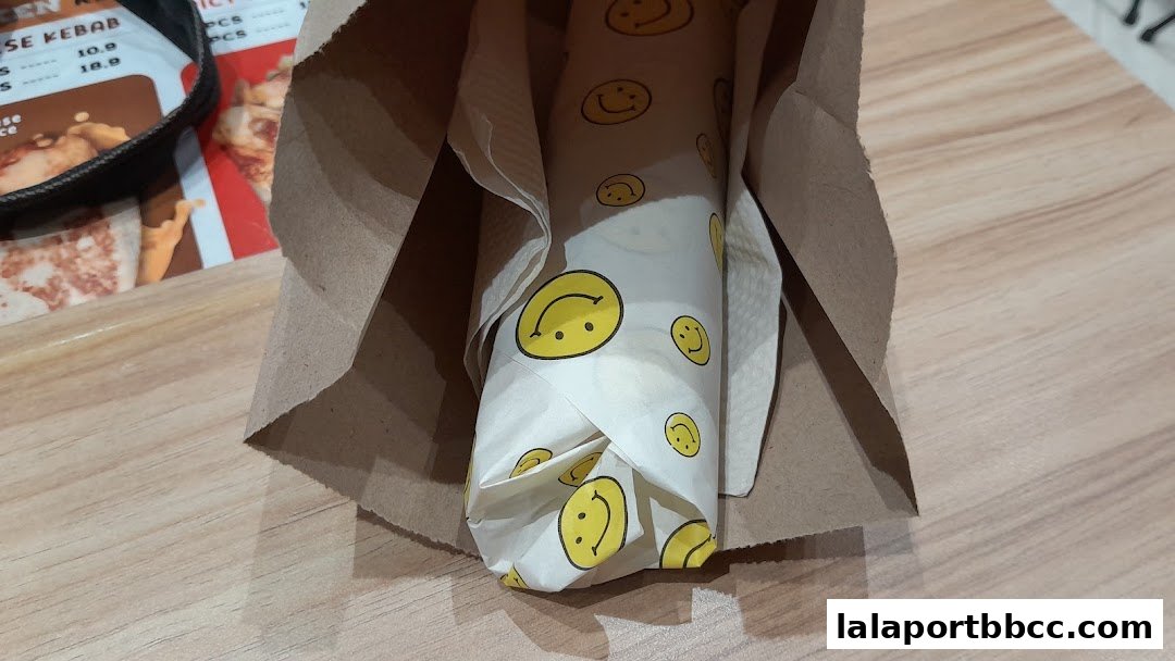 A kebab wrapped in smiley face paper sits in a brown paper bag.