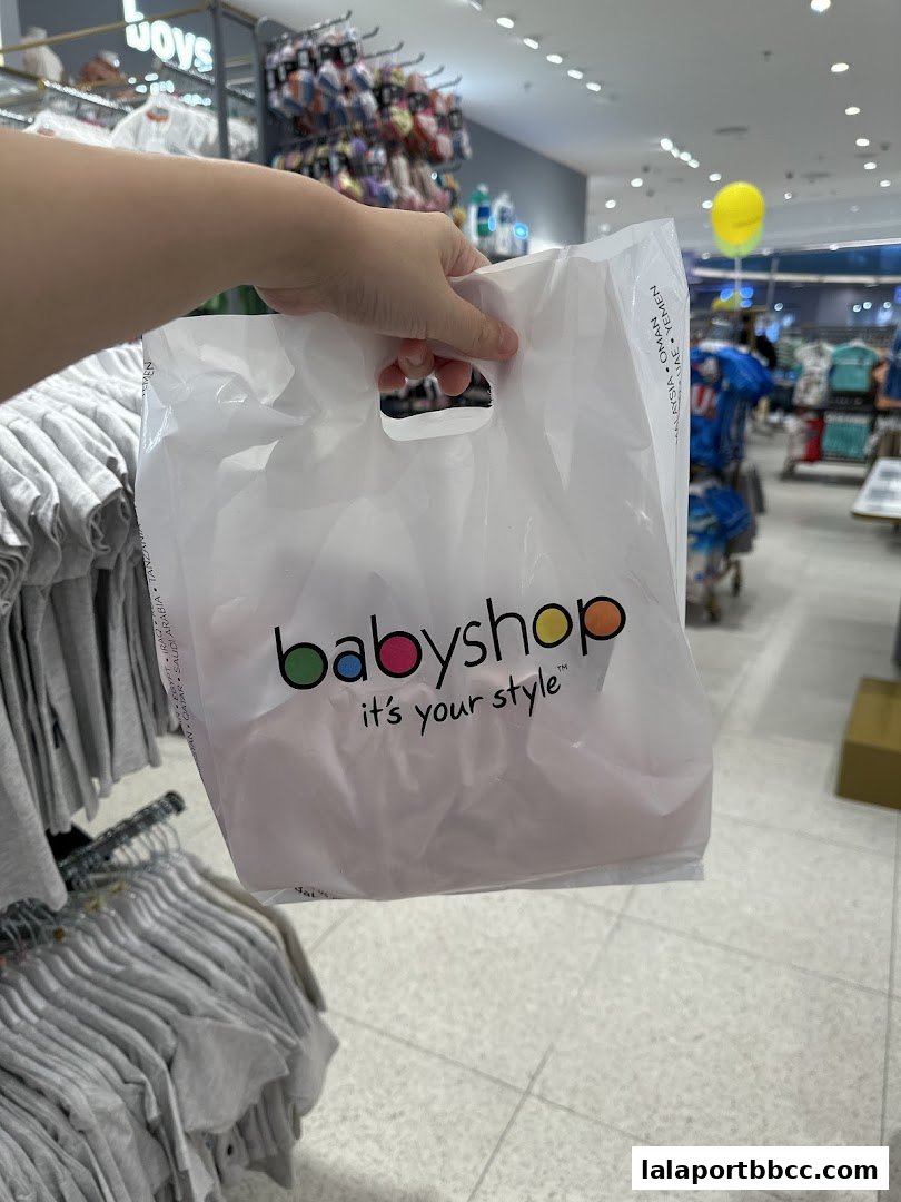 A hand holds a Babyshop shopping bag inside a store with clothing racks.