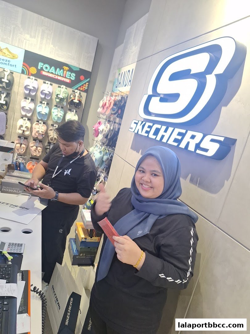 A woman gives a thumbs up in a Skechers store, with footwear displays and staff visible.