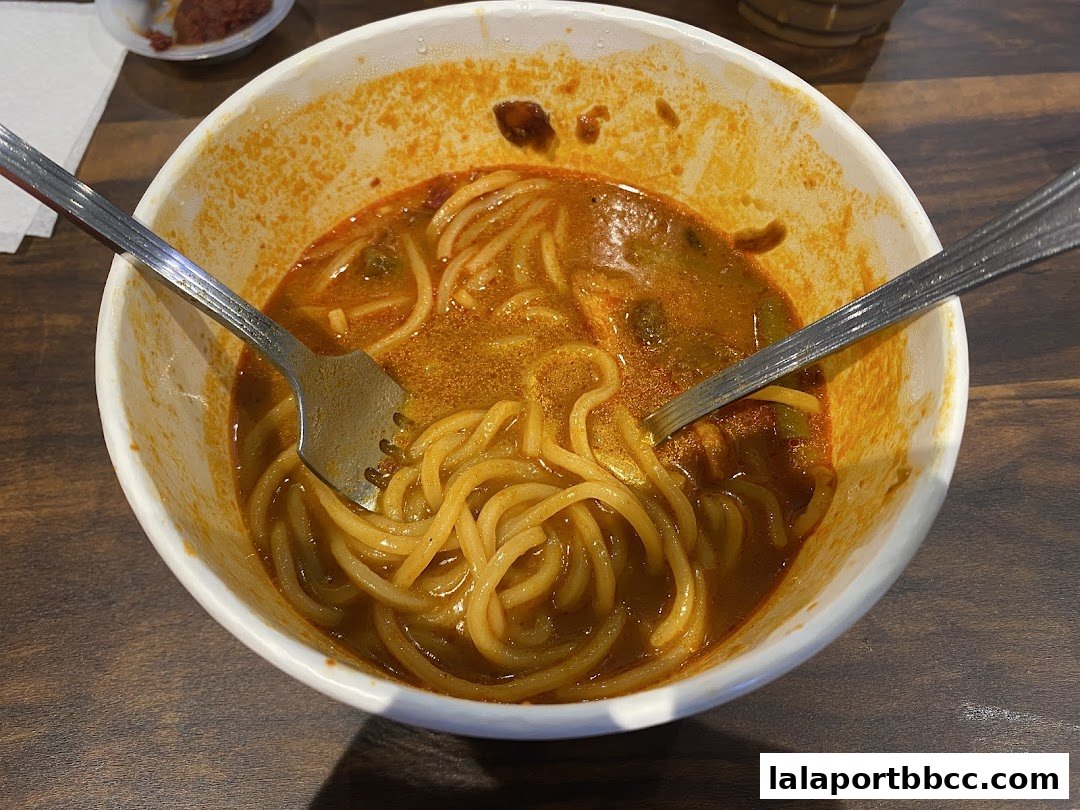 A bowl of noodles in a rich, orange broth with two forks, ready to be eaten.