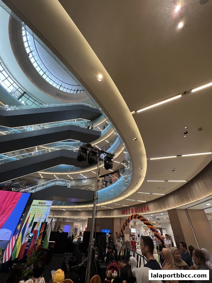A wide shot of a shopping mall interior with escalators, a stage, and a crowd of people.
