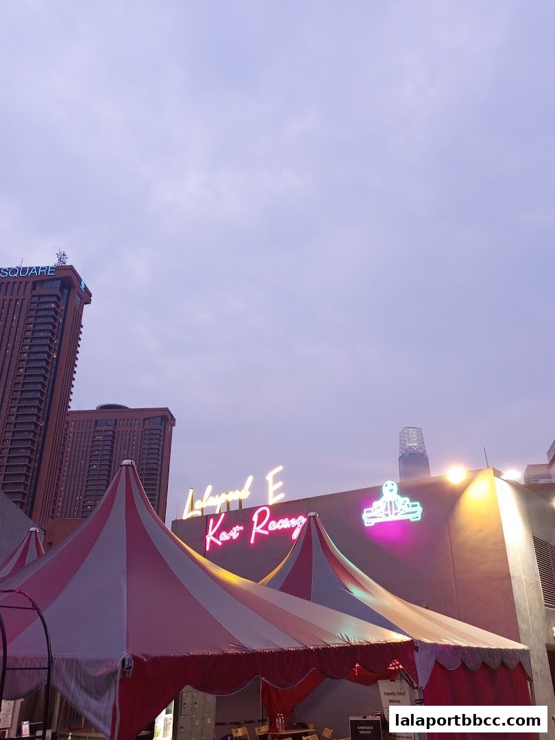 Neon sign for Lalaspeed E Kart Raceway above tents with city buildings in the background.