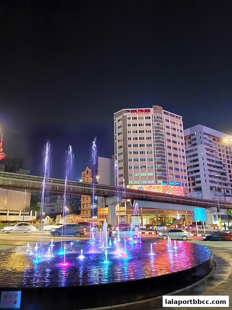 A vibrant fountain with colorful lights at night, with buildings and traffic in the background.