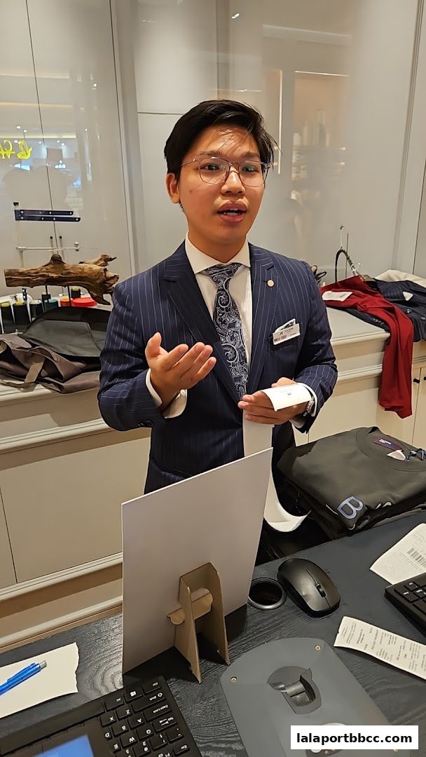 A man in a pinstripe suit stands behind a counter, gesturing while holding a receipt.