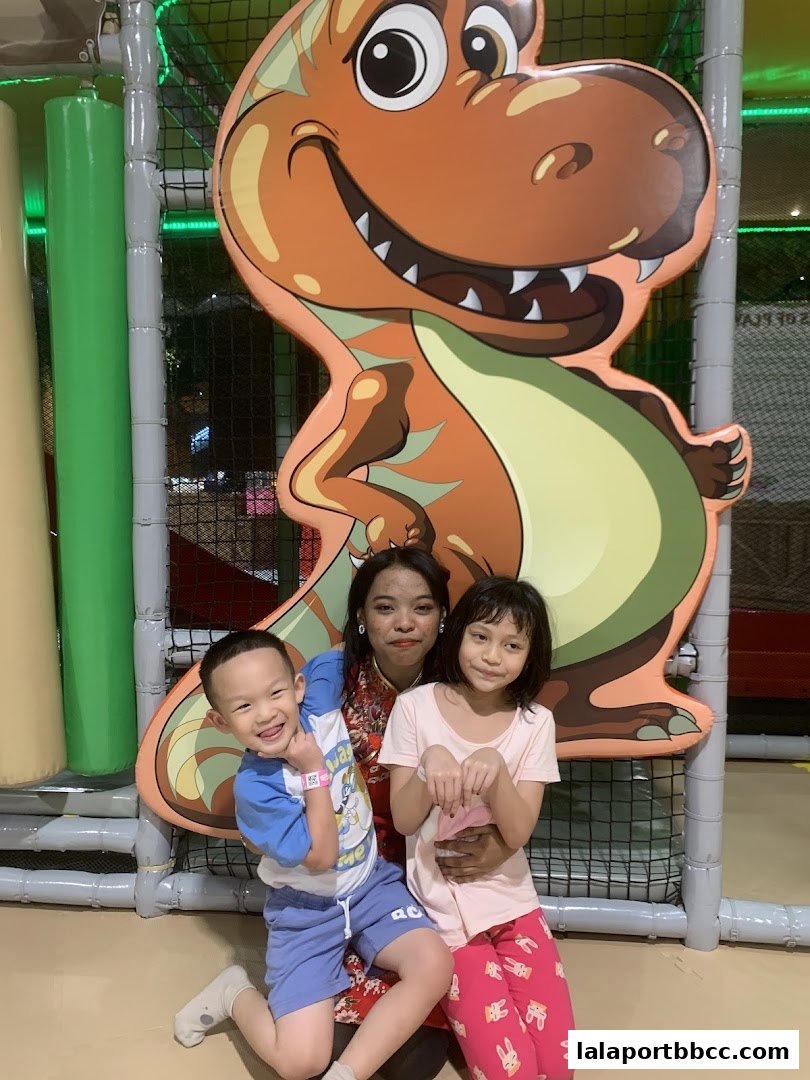 A woman and two children pose in front of a large dinosaur cutout at an indoor playground.