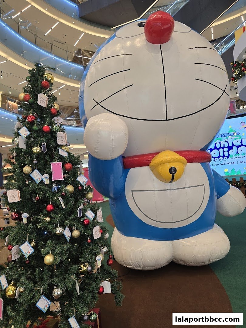A giant inflatable Doraemon character stands next to a decorated Christmas tree in a mall.
