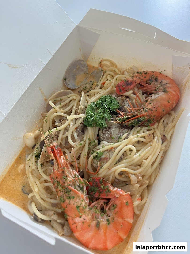 A close-up of a seafood pasta dish in a takeout box, featuring large prawns and clams.