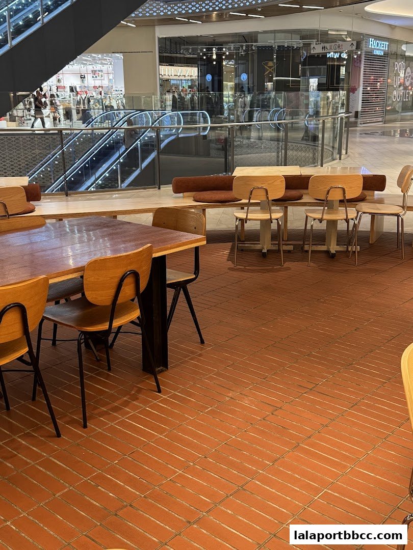 Interior view of a cafe with tables and chairs, overlooking a shopping mall with escalators.