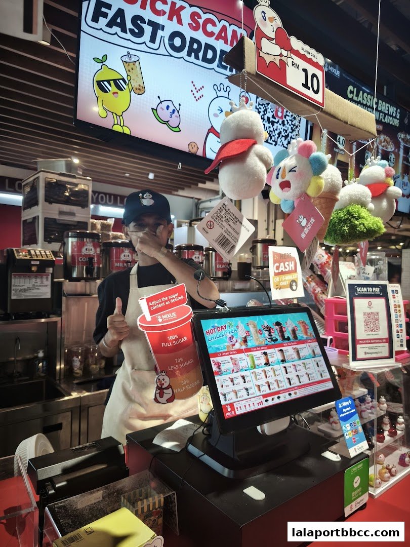A MIXUE employee stands behind the counter with a digital menu and promotional items.