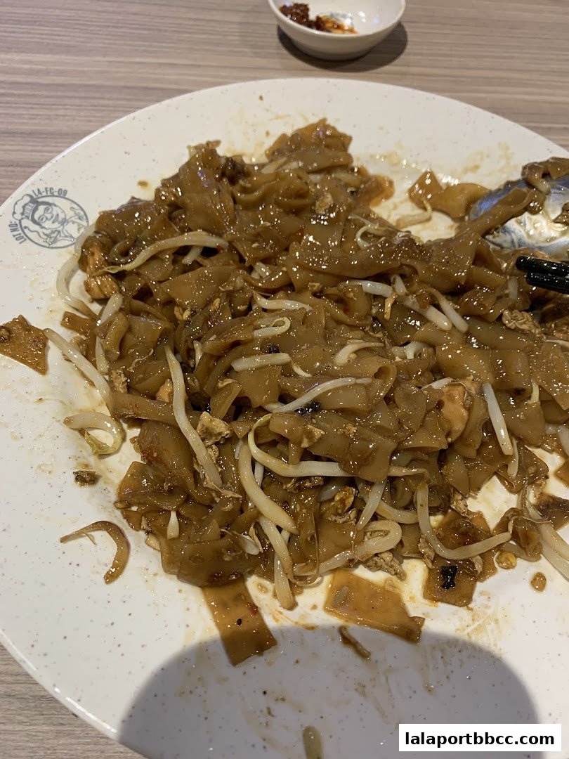 A plate of Hokkien Mee noodles with bean sprouts and a small bowl of chili sauce.