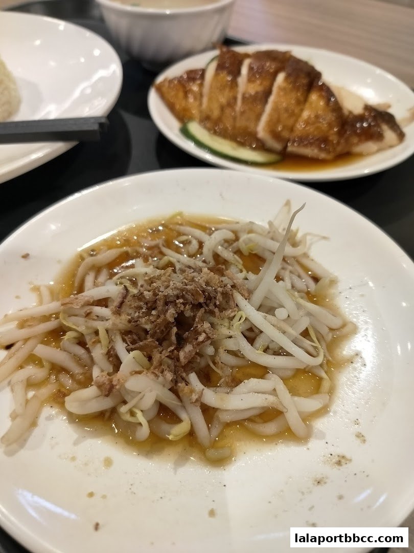 A plate of bean sprouts with sauce and fried shallots, next to chicken rice and soup.
