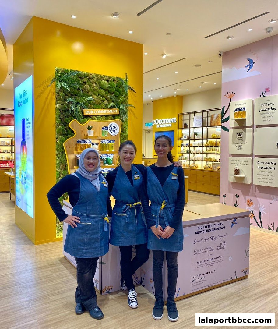 Three smiling staff members stand in front of a L'Occitane store display featuring natural elements and marketing information.