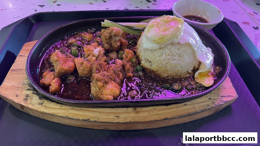 A sizzling claypot dish with fried chicken, rice, a fried egg, and sauce.