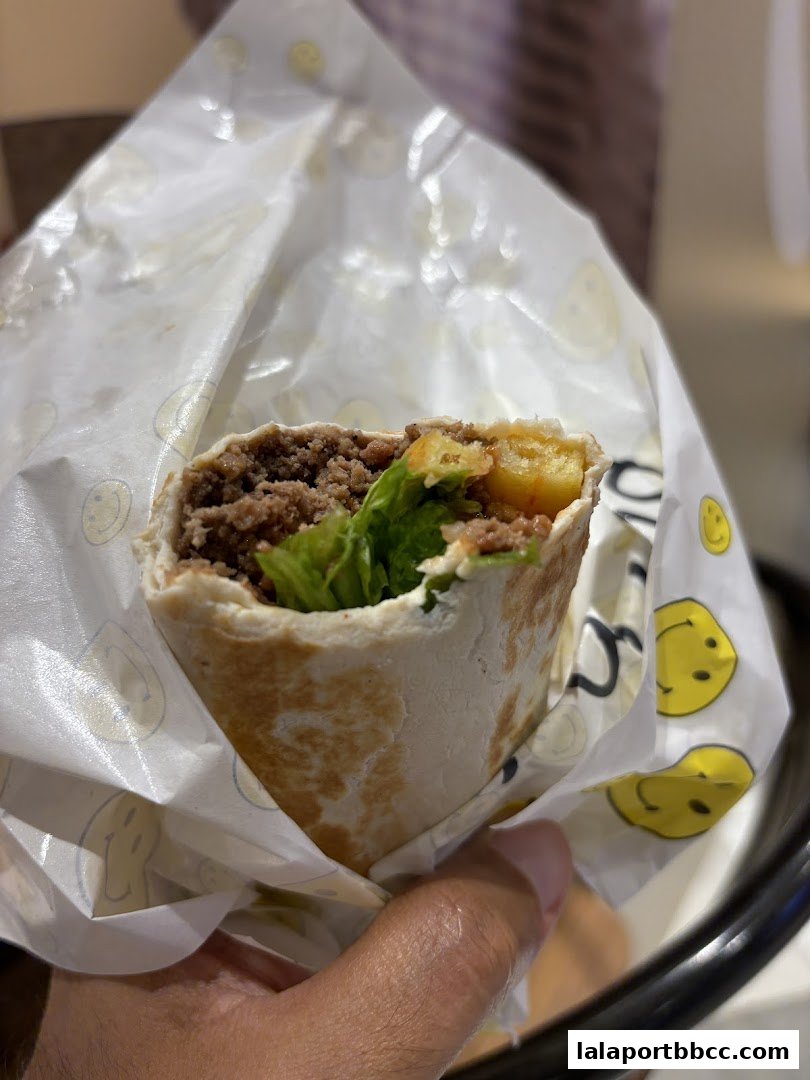 A hand holds a freshly wrapped kebab filled with meat, lettuce, and fries.