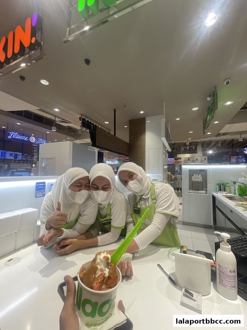Three smiling staff members in green aprons pose with a frozen yogurt dessert.