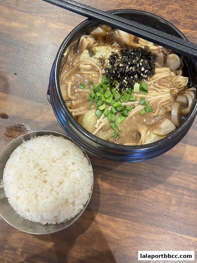 A steaming bowl of Korean stew with enoki mushrooms, seaweed, and green onions, served with a side of rice.