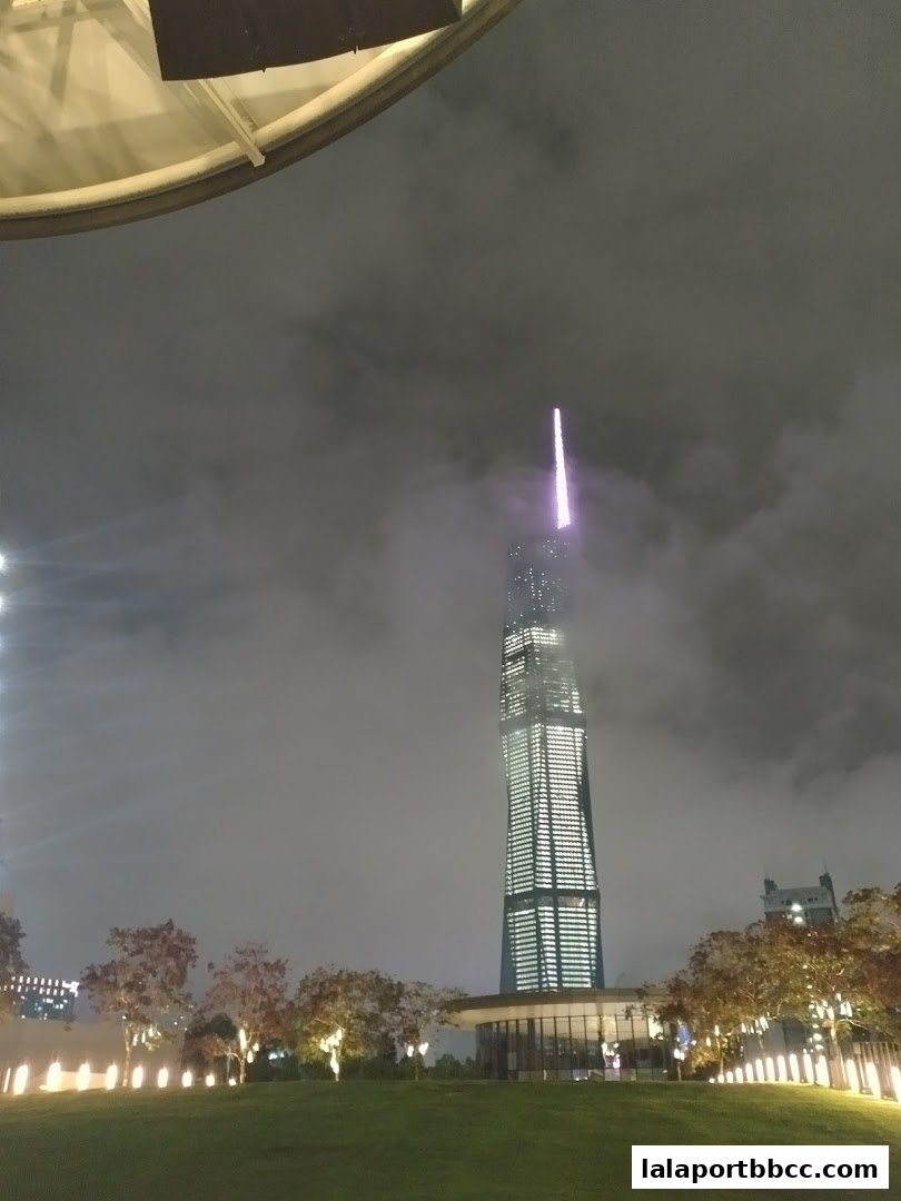 A tall, illuminated skyscraper pierces the cloudy night sky, with trees and lights in the foreground.