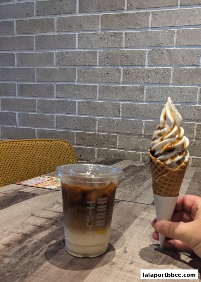 A hand holds a soft-serve ice cream cone next to a iced coffee drink on a table.