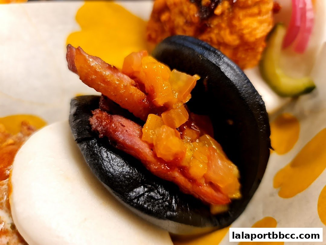 A close-up of a black bao bun filled with meat and toppings, with other food items in the background.