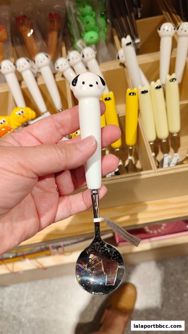 A hand holds a cute dog-themed spoon with various novelty utensils displayed in the background.