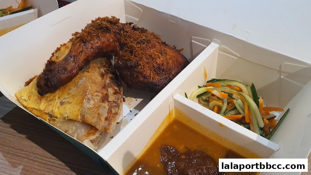 A takeaway box filled with a crispy fried chicken leg, roti canai, salad, and curry.