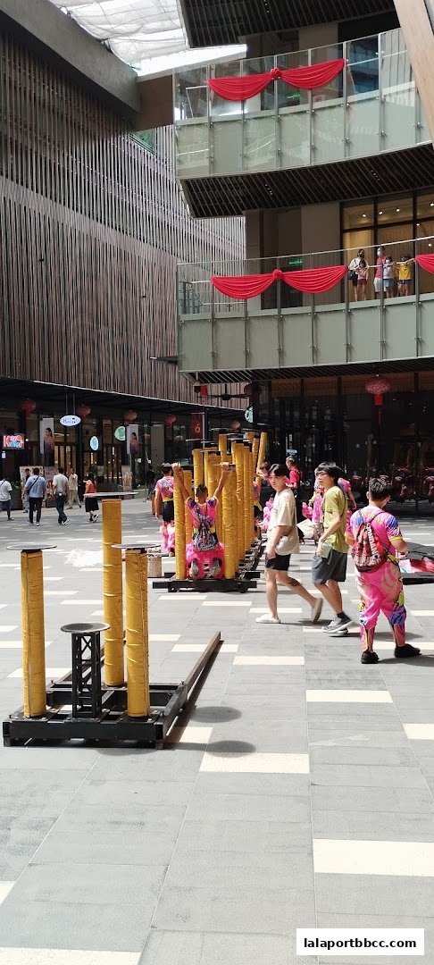 People in colorful outfits perform near bamboo poles in a modern outdoor plaza.