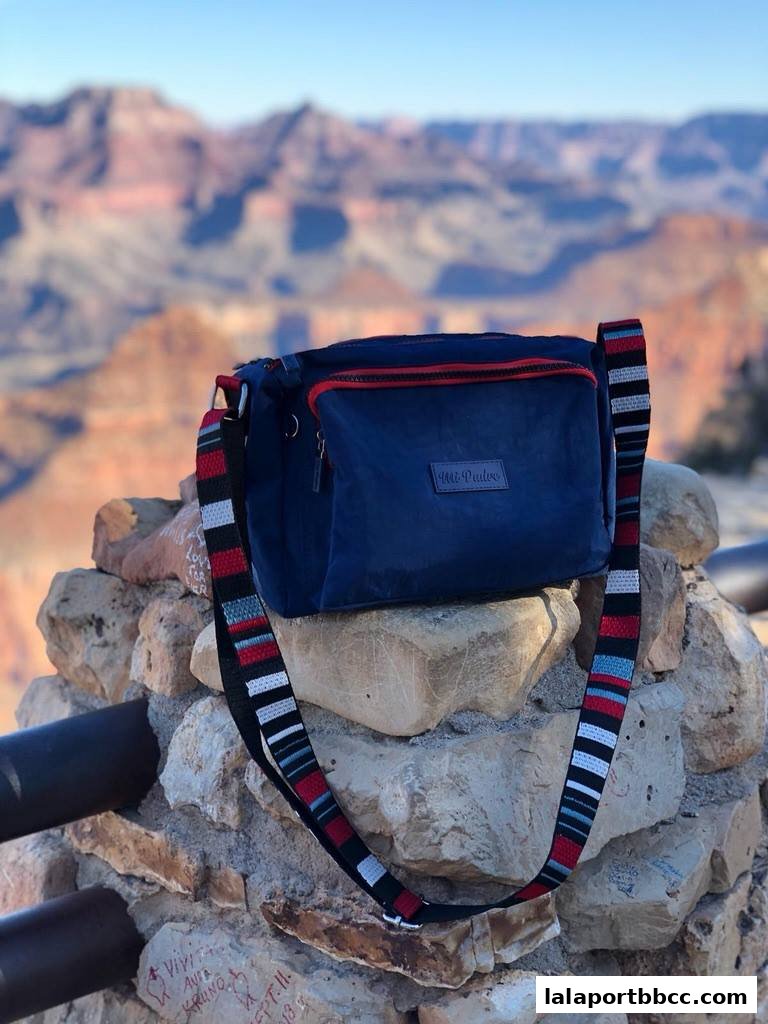 A blue crossbody bag with a striped strap rests on rocks with a canyon in the background.