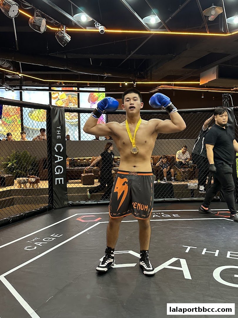 A young man in boxing gloves and shorts poses victoriously in a fighting cage.