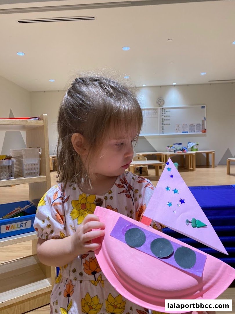 A young child proudly holds a handmade craft project featuring a pink paper plate and a triangular shape.