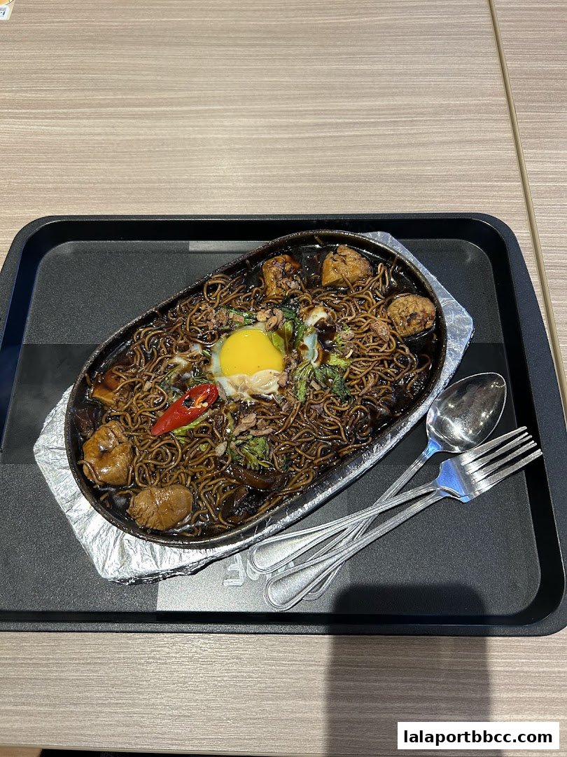 A sizzling plate of noodles with a fried egg, meat, and vegetables, served with cutlery.