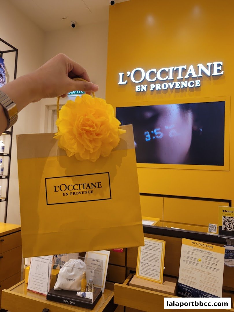A hand holds a L'Occitane shopping bag with a yellow flower decoration in front of the store.