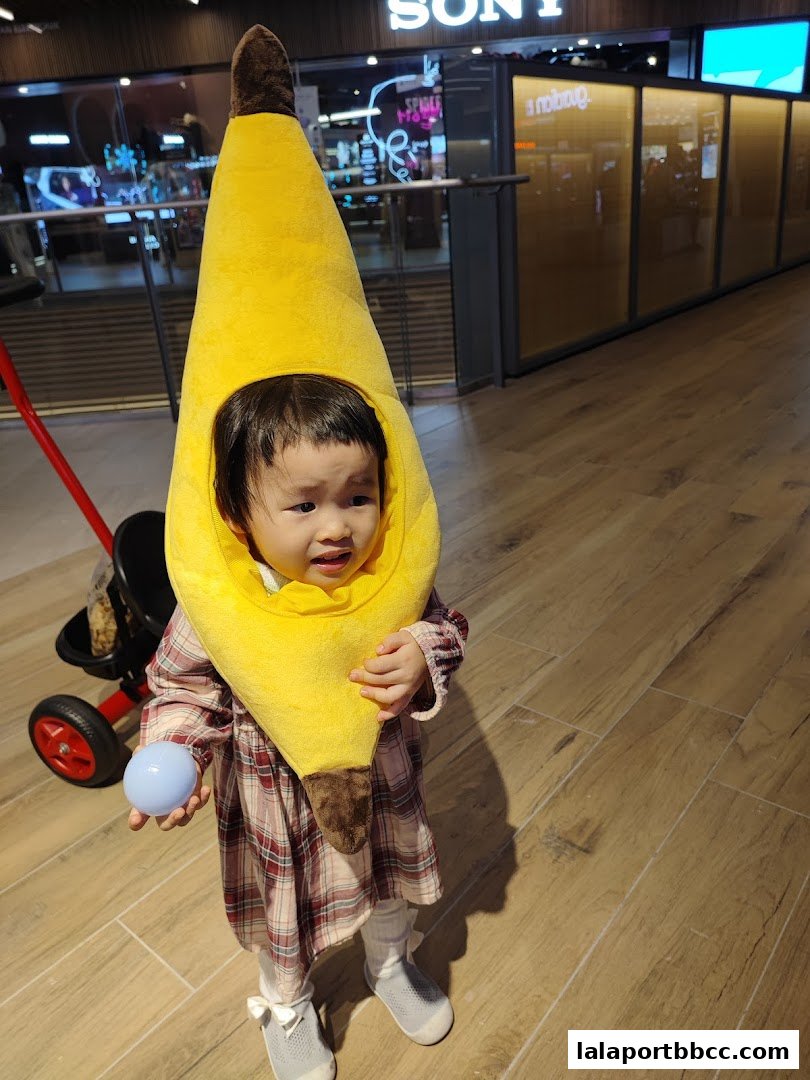 A child in a banana costume stands in a mall hallway, holding a blue ball.
