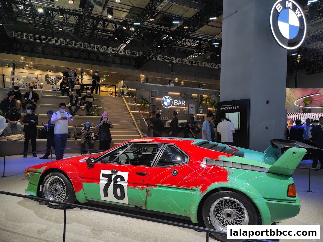 A colorful BMW M1 car is displayed at an event with people watching and a bar in the background.