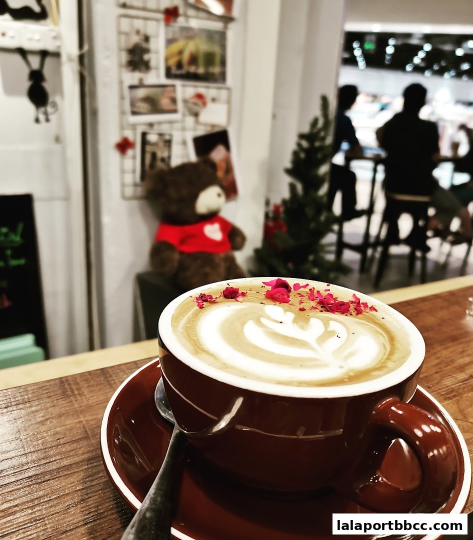 A close-up of a latte with latte art and flower petals, with a teddy bear and people in the background.