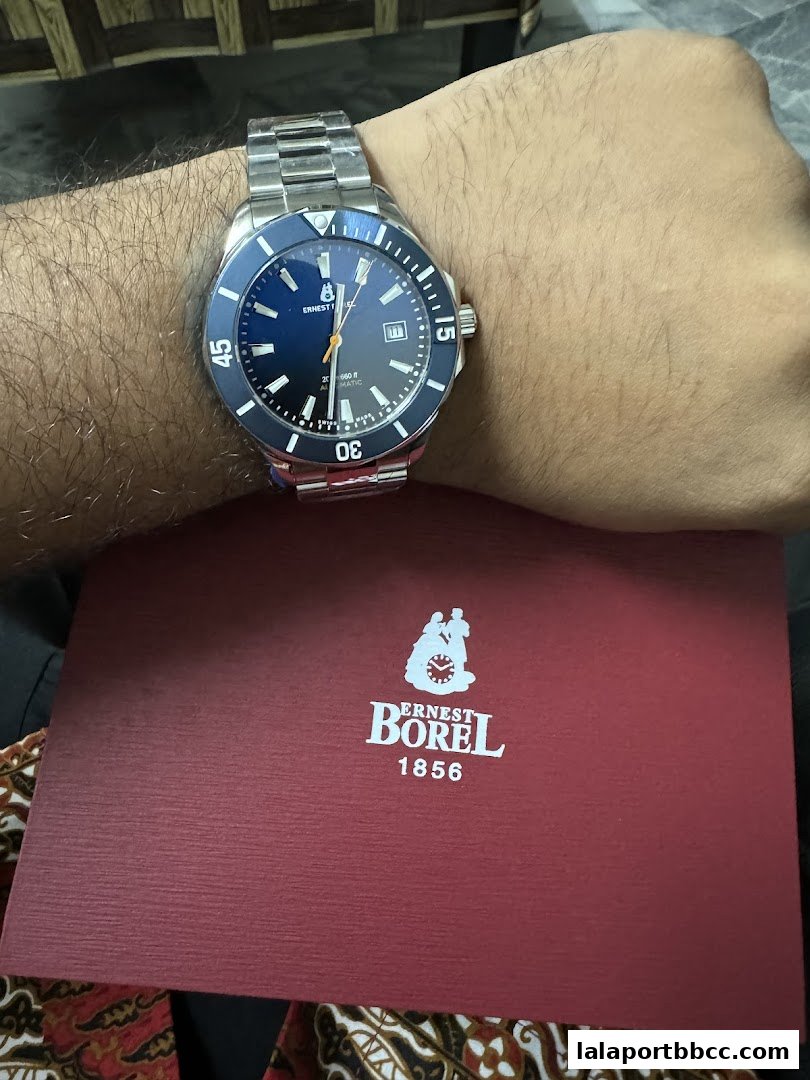 A person wearing an Ernest Borel watch with a blue dial and a red presentation box.