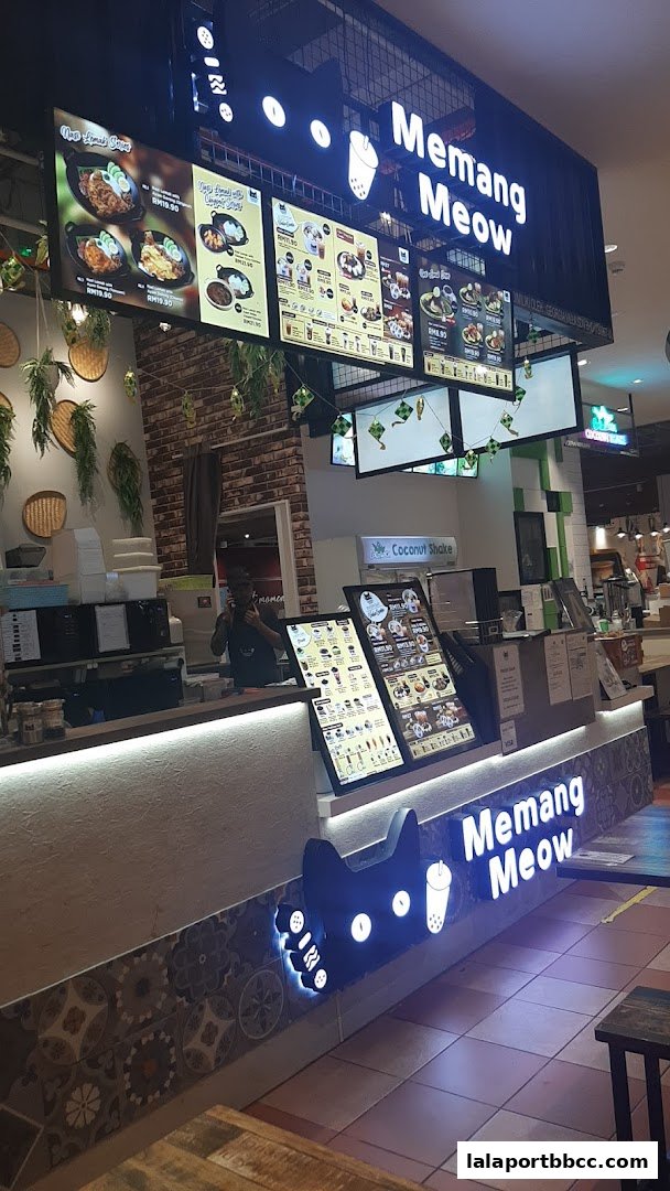 A brightly lit food stall with a prominent 'Memang Meow' sign and digital menus displaying food items.
