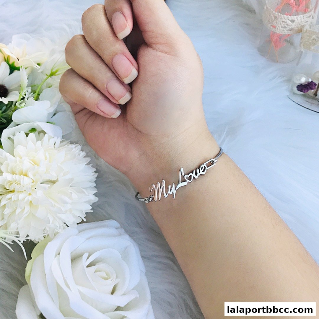 A silver bracelet with 'My Love' inscribed is worn on a woman's wrist, surrounded by white flowers.