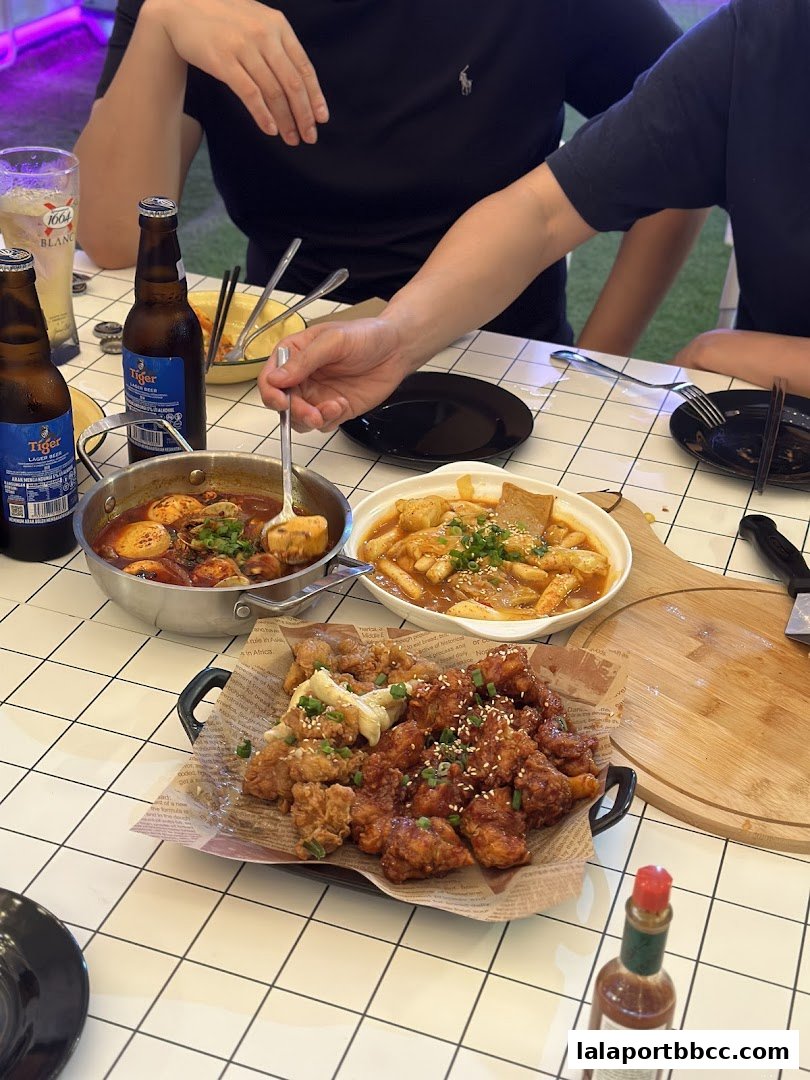 A table laden with Korean dishes, including fried chicken, tteokbokki, and a stew, with bottles of Tiger beer.