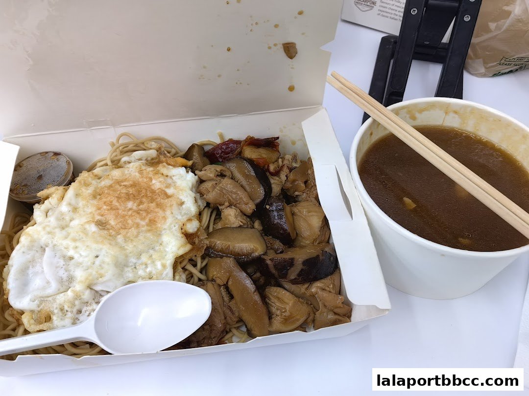 A takeaway box filled with noodles, a fried egg, and meat, served with a side of soup.